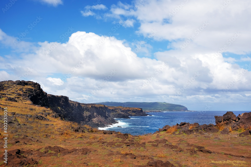 View of the coast of Easter Island, covered by red volcanic dust and green vegetation, against a blue sky with white clouds.