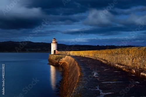 An empty promenade to the old lighthouse at night, close-up. Rocky shores in the background. Dramatic cloudscape. Ardrishaig, Crinan canal, Scotland, UK. Travel destinations, sightseeing, landmarks photo
