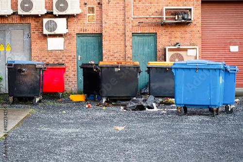 Back yard of an office building with colored rubbish bins, fire doors and utility room entrance. Many air conditioner units mounted on a brick wall. Dirt on the ground