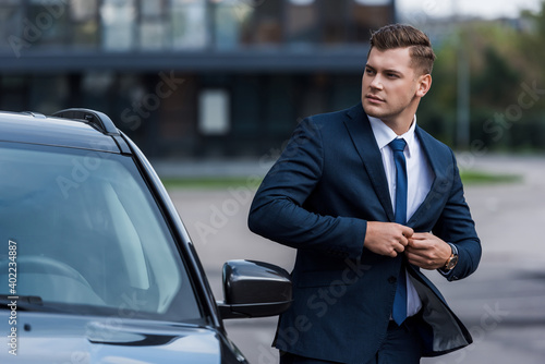 businessman buttoning up blazer near car, blurred foreground. © LIGHTFIELD STUDIOS