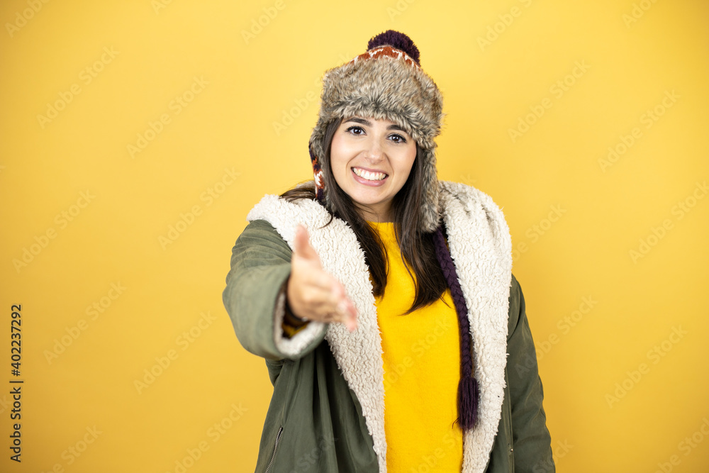 Young beautiful woman wearing a hat and a green winter coat over yellow background smiling friendly offering handshake as greeting and welcoming Young beautiful woman wearing a hat and a green winter coat over yellow background smiling friendly offering handshake as greeting and welcoming
