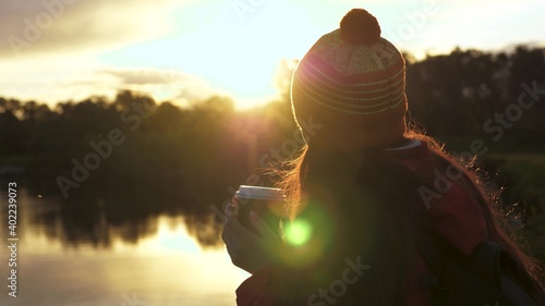 A tourist drinks tea from a mug in sun. Woman Traveler holds mug with hot coffee in her hands and looks at sunset byriver. Free traveler girl admiring scenery. Hiker at rest drinking hot cocoa