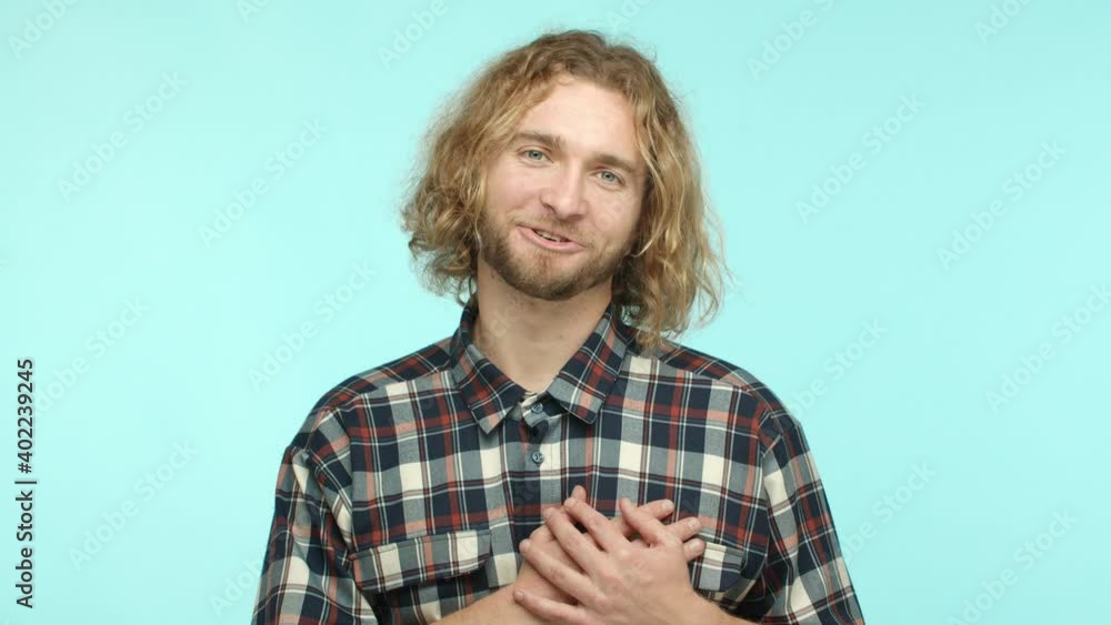 Slow motion of smiling handsome blond man feeling touched and flattered, holding hands on heart and sighing, looking at camera and saying thank you, standing over turquoise background Slow motion of smiling handsome blond man feeling touched and flattered, holding hands on heart and sighing, looking at camera and saying thank you, standing over turquoise background