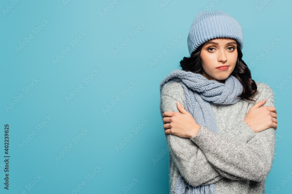  brunette woman in hat and scarf hugging herself while looking at camera isolated on blue