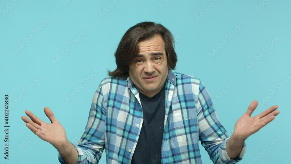 Slow motion of reluctant middle-aged guy shrugging and grimacing, raising hands up and looking clueless, know nothing, standing indecisive against blue background Slow motion of reluctant middle-aged guy shrugging and grimacing, raising hands up and looking clueless, know nothing, standing indecisive against blue background
