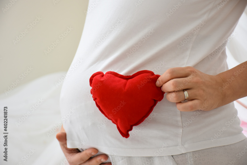 Pregnant woman holding a red heart sign. Pregnant woman put a toy heart to her stomach on white background Pregnant woman holding a red heart sign. Pregnant woman put a toy heart to her stomach on white background