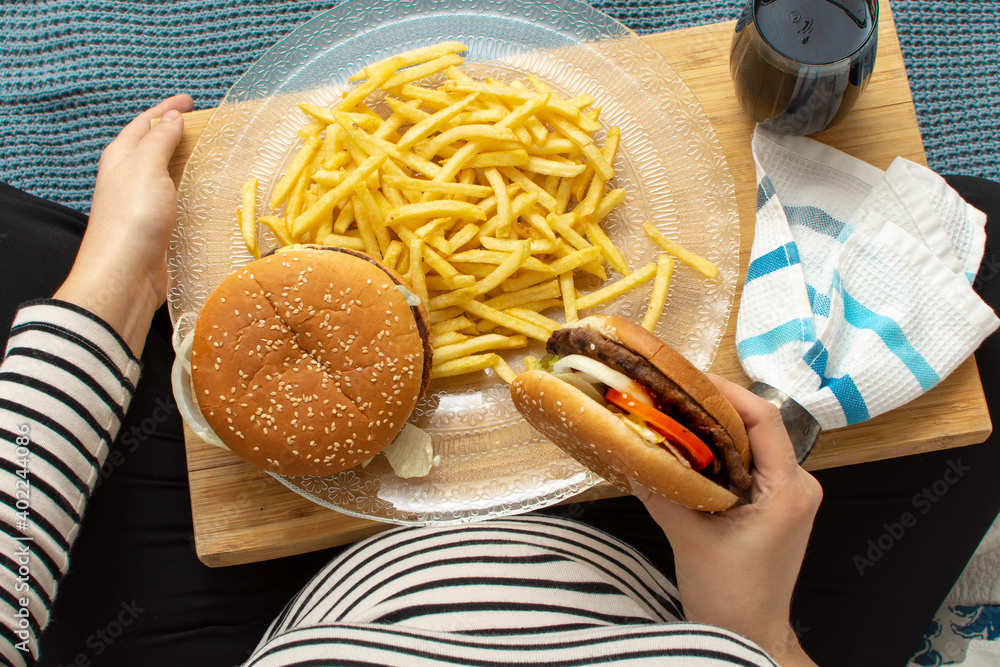 © Ersin - Cropped image of young hungry pregnant woman eating a hamburger and fried potatoes. Concept of unhealthy nutrition during pregnancy, Mother waiting baby. Close-up of belly and junk food (fast food).