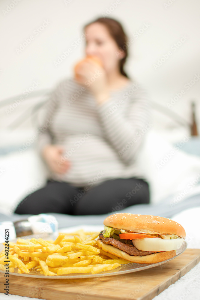 young hungry pregnant woman eating a hamburger and fried potatoes. Concept of unhealthy nutrition during pregnancy, Mother waiting baby. Close-up of belly and junk food (fast food). young hungry pregnant woman eating a hamburger and fried potatoes. Concept of unhealthy nutrition during pregnancy, Mother waiting baby. Close-up of belly and junk food (fast food).