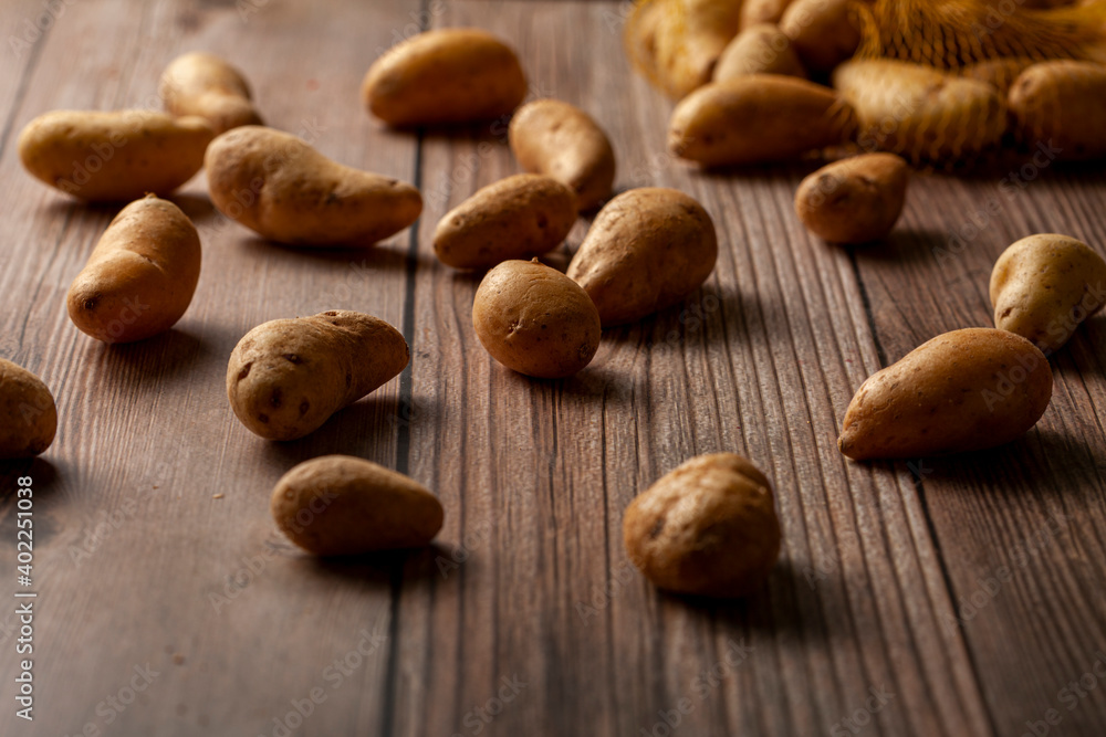Shallow depth of field dark image of small irregular shaped ratte potatoes scattered on the surface of a wooden table from a tipped over mesh sack. An artistic versatile low light still life photo. Shallow depth of field dark image of small irregular shaped ratte potatoes scattered on the surface of a wooden table from a tipped over mesh sack. An artistic versatile low light still life photo.