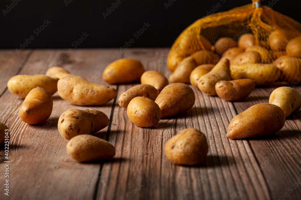 Shallow depth of field dark image of small irregular shaped ratte potatoes scattered on the surface of a wooden table from a tipped over mesh sack. An artistic versatile low light still life photo. Shallow depth of field dark image of small irregular shaped ratte potatoes scattered on the surface of a wooden table from a tipped over mesh sack. An artistic versatile low light still life photo.