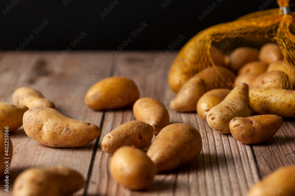 Shallow depth of field dark image of small irregular shaped ratte potatoes scattered on the surface of a wooden table from a tipped over mesh sack. An artistic versatile low light still life photo. Shallow depth of field dark image of small irregular shaped ratte potatoes scattered on the surface of a wooden table from a tipped over mesh sack. An artistic versatile low light still life photo.
