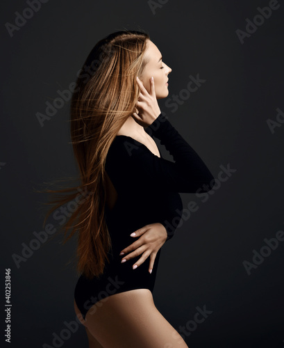 Young sensual woman in black bodysuit stands side to camera with closed of pleasure eyes holding strand brushing her long silky hair over dark background. Haircare, beauty, wellness, hairstyle concept