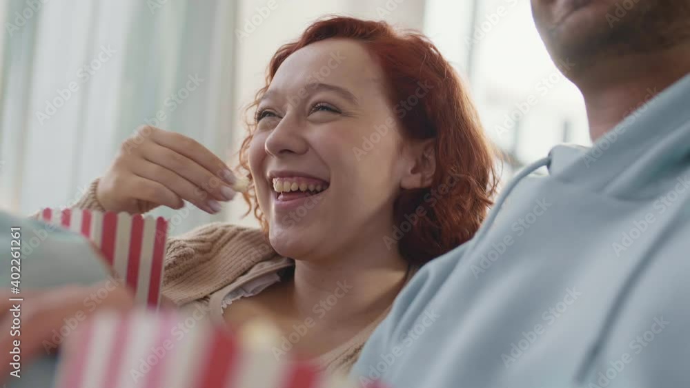 Low angle close-up of young Caucasian woman sitting next to unrecognizable person indoors, eating popcorn from red and white striped paper box, laughing Low angle close-up of young Caucasian woman sitting next to unrecognizable person indoors, eating popcorn from red and white striped paper box, laughing
