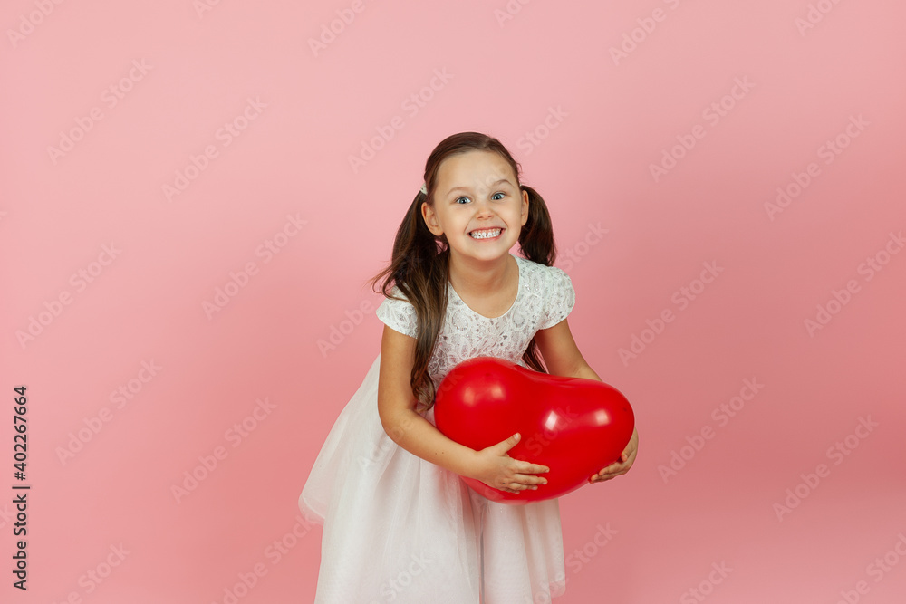 delighted, pleased girl with ponytails and in a white dress stands half-sideways and holds a red balloon in the form of a heart, isolated on a pink background. delighted, pleased girl with ponytails and in a white dress stands half-sideways and holds a red balloon in the form of a heart, isolated on a pink background.