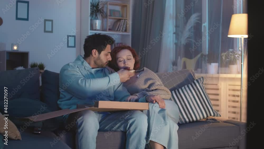 Medium long steadicam of young Mixed-Race man eating pizza from box, sharing his slice with red-haired Caucasian girlfriend sitting near on couch in living room. Couple watching invisible TV at night Medium long steadicam of young Mixed-Race man eating pizza from box, sharing his slice with red-haired Caucasian girlfriend sitting near on couch in living room. Couple watching invisible TV at night