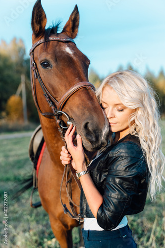A pretty blonde in a black jacket stands in a field next to a horse at sunset. © cinematri
