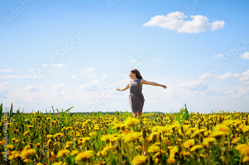 Beautiful young woman on a field with green grass and yellow dandelion flowers in a sunny day. Girl on nature with yellow flowers and blue sky.