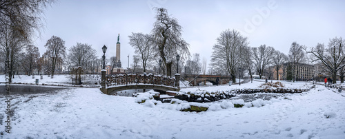 Panoramic winter landscape in snowy park with beautiful bridge over small pond, street light and covered in snow tress. View of Bastion Hill park and The Freedom Monument in Riga, Latvia © Maria Vonotna