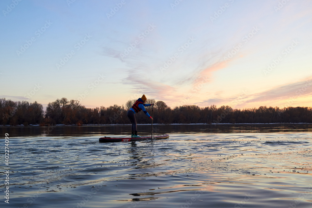 Silhouette of young girl rowing on stand up paddle board (SUP) on Danube river at winter sunset. Backlight