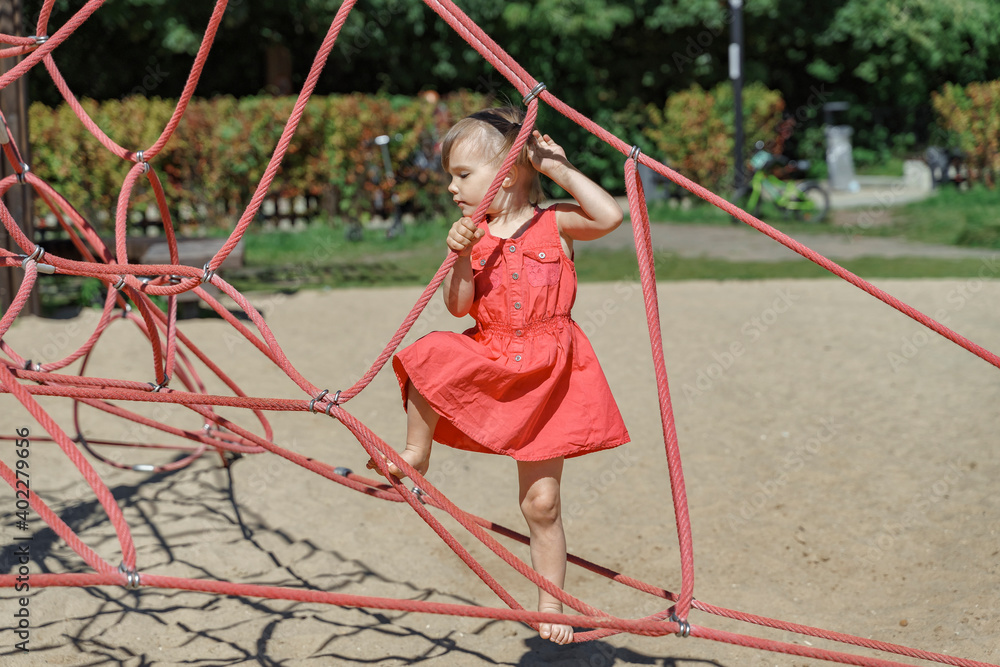 beautiful little three-year-old child in a red dress in a rope web on the playground