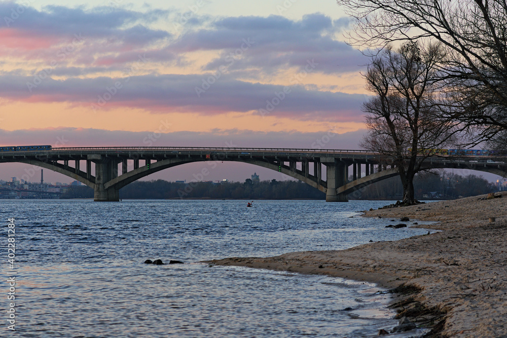Lonely bare tree on the bank of the Dnieper River. Metro (subway) Bridge at the background. Winter sunset landscape photo. Kyiv, Ukraine Lonely bare tree on the bank of the Dnieper River. Metro (subway) Bridge at the background. Winter sunset landscape photo. Kyiv, Ukraine