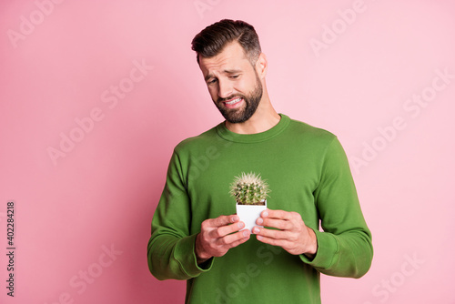Portrait of attractive dissatisfied guy holding in hands potted cactus oops isolated over pink pastel color background