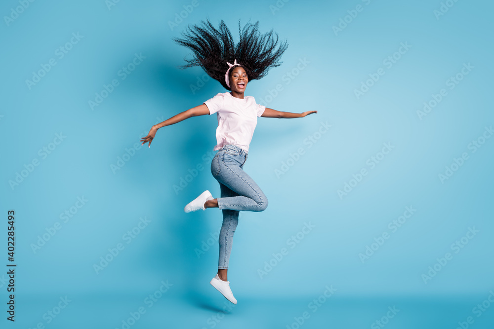 Full length photo of shiny pretty afro woman wear pink t-shirt jumping high one leg hands arms sides isolated blue color background