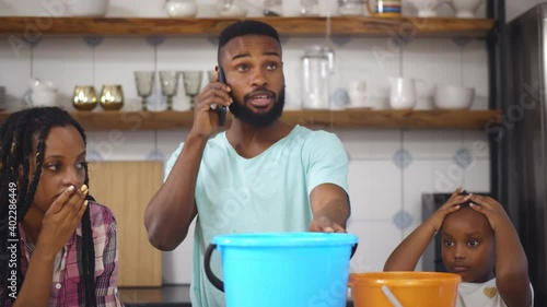 Worried african family collecting water leaking from ceiling while father calling plumber at home