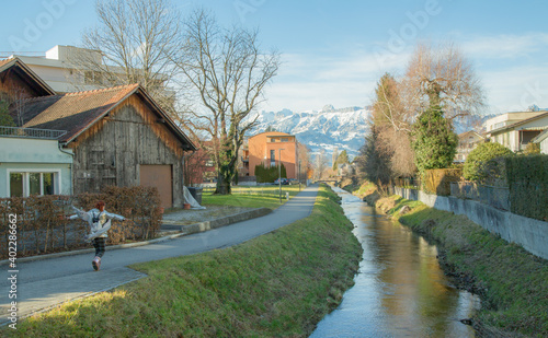 Happy girl in Alps mountains in Liechtenstein. Medieval Red House, calm narrow mountain river and jogging track, residential buildings, blue sky and snow-capped mountains. Liechtenstein, Vaduz