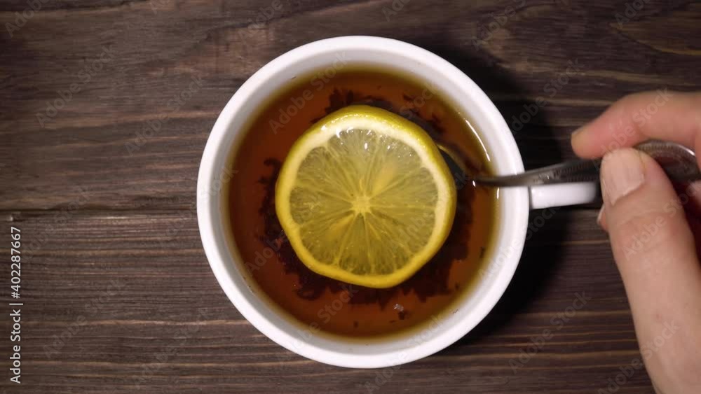 Female hand stirring tea with lemon in a white cup with a spoon. Tea on a table from dark boards. Close-up. Female hand stirring tea with lemon in a white cup with a spoon. Tea on a table from dark boards. Close-up.