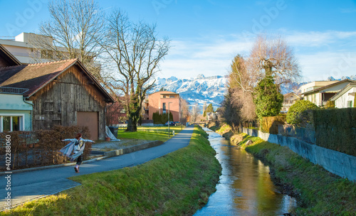 Happy girl in Alps mountains in Liechtenstein. Medieval Red House, calm narrow mountain river and jogging track, residential buildings, blue sky and snow-capped mountains. Liechtenstein, Vaduz