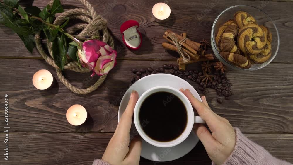 Female hands on a white cup of coffee. Table decoration for Valentine's Day.A romantically decorated table with candles, a rose flower and a gold ring in a box. Female hands on a white cup of coffee. Table decoration for Valentine's Day.A romantically decorated table with candles, a rose flower and a gold ring in a box.
