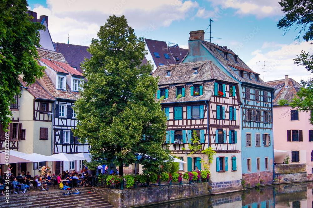 Fototapeta premium Colorful half-timbered houses and a restaurant with an outdoor area at a canal in the historical city of Strasbourg. The house facades reflect in the water.