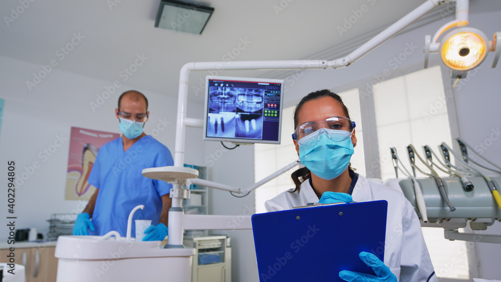 Obraz premium Dentistry doctor interrogating patient and taking notes on clipboard before surgery. Doctor and nurse working in modern orthodontic office, writing and examining person wearing protection mask