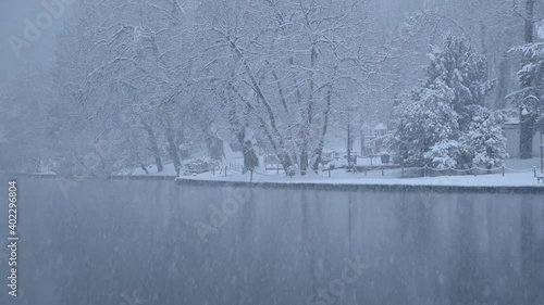 Snow falling on lake Bled shore, Slovenia. Snowstorm in winter season. Calm view of snowflakes falling on ground. Static shot, wide angle, real time