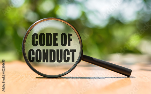 Magnifying glass with text CODE OF CONDUCT on wooden table and green background.