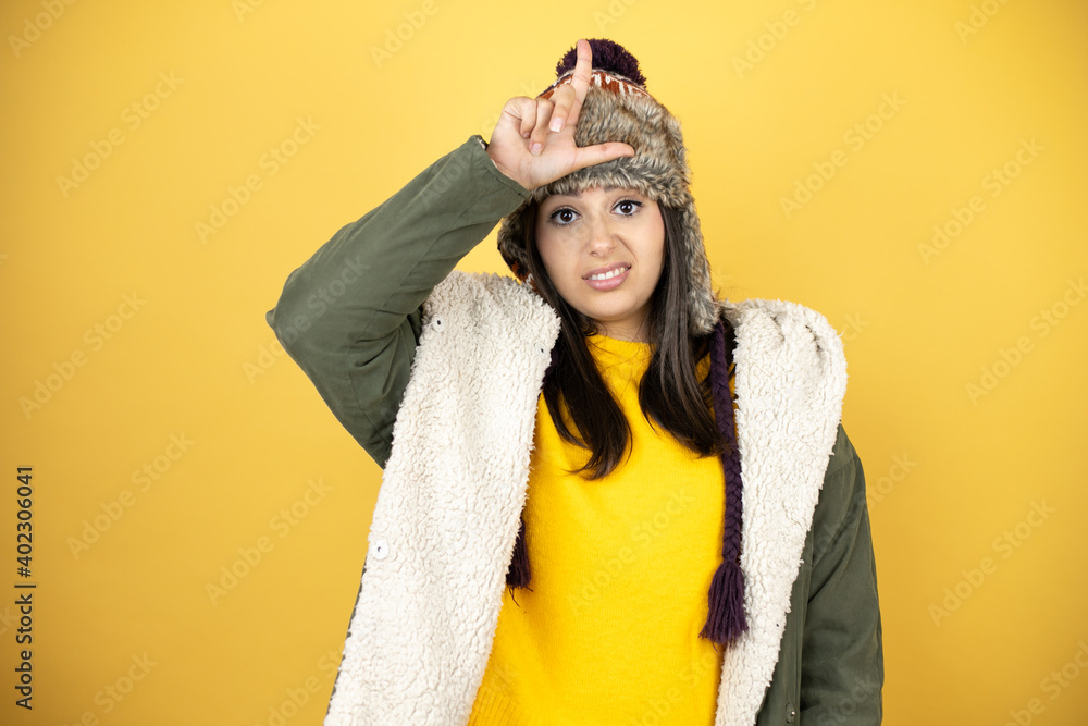 Young beautiful woman wearing a hat and a green winter coat over yellow background making fun of people with fingers on forehead doing loser gesture mocking and insulting.
