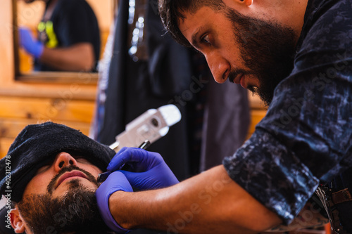 Close-up of a Latin barber cutting the beard of a bearded man - Barber with blue gloves shaves a man's beard with a razor in the living room