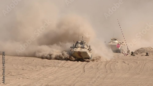 A column of armored vehicles passes through a checkpoint to perform a combat mission in a military conflict zone, dust rises from under the wheels.