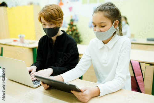 Students in medical masks sit at the table and take part in an online lesson using gadgets
