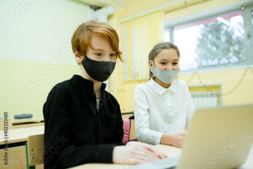 A girl in a white shirt and a boy in a black sweater in protective equipment study online using a computer