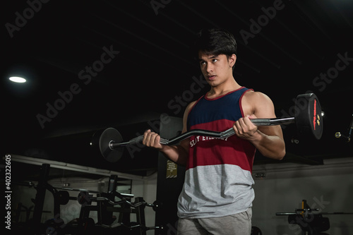 A young asian man of intermediate muscle development doing some EZ barbell curls at the gym.