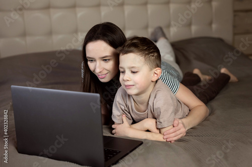 mom and son are looking at the computer screen. the models are located on a bed in a wooden house. Beige tones.