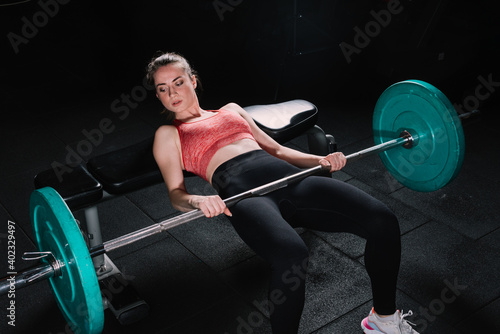 Young beautiful woman doing hip thrust. She poses with barbells in the gym.