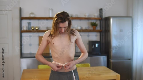 Young thin shirtless man measuring waist with tape at home