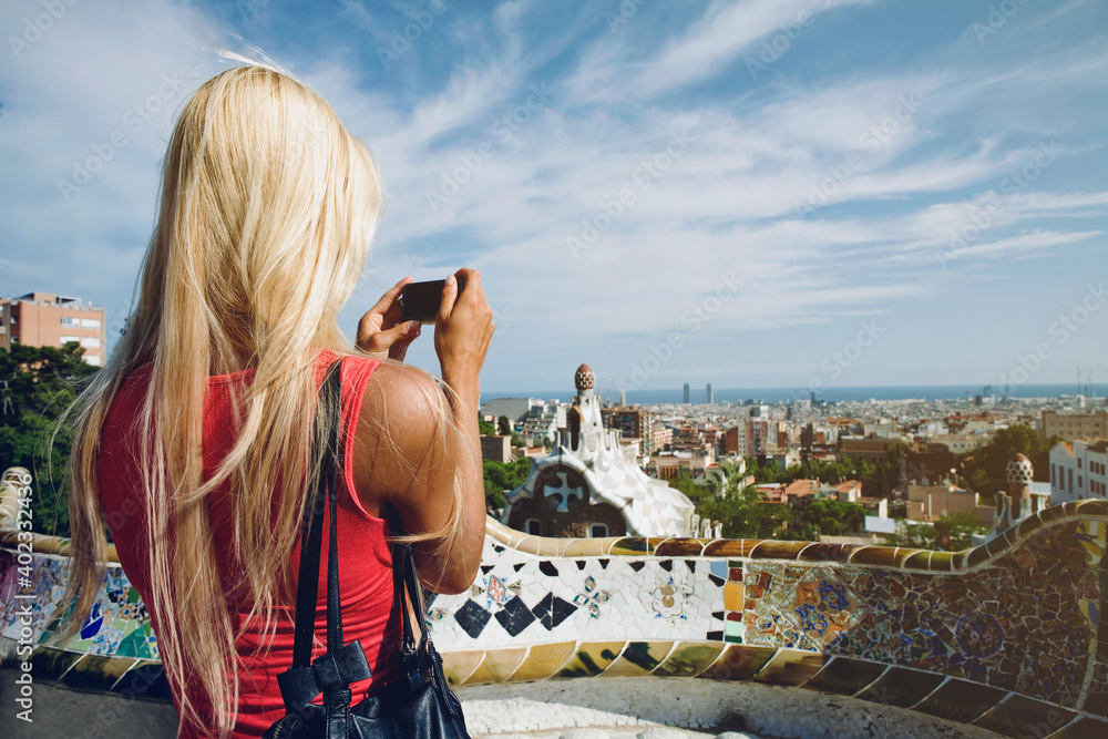 Blondy caucasian Girl taking picture of Barcelona from the famous terrace of of Park Guell, architectural landmark designed by the famous architect Antonio Gaudi.