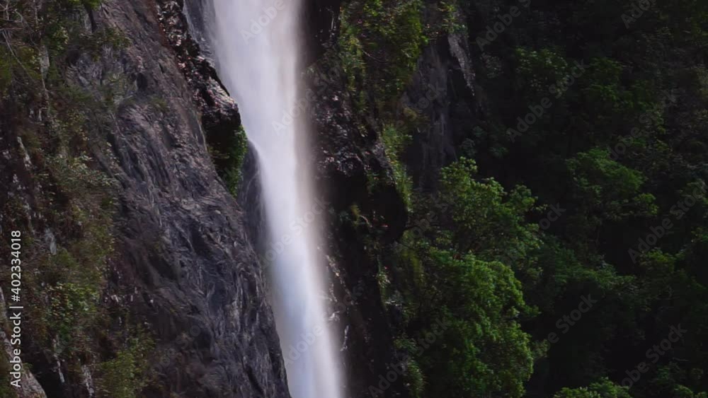 View of a water falling down the rocky wall with lush green bushes in the background, dark and moody morning. View of a water falling down the rocky wall with lush green bushes in the background, dark and moody morning.