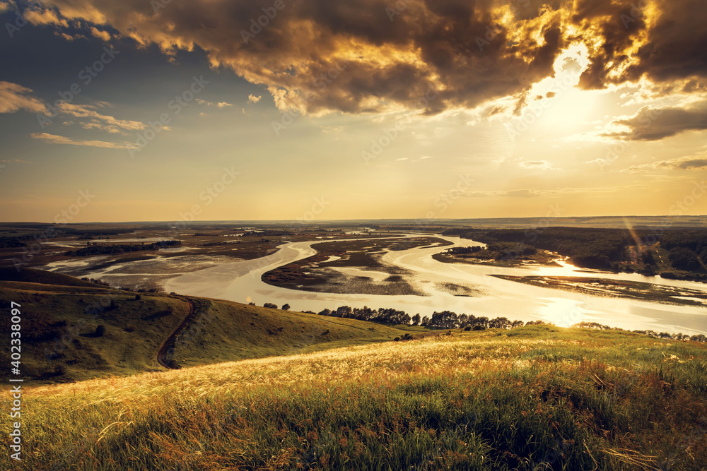 Fototapeta premium Sunny summer landscape with river, fields, green hills and beautiful clouds in blue sky. Pretty view of summer scenery on a nice day. Green trees growing along the river.