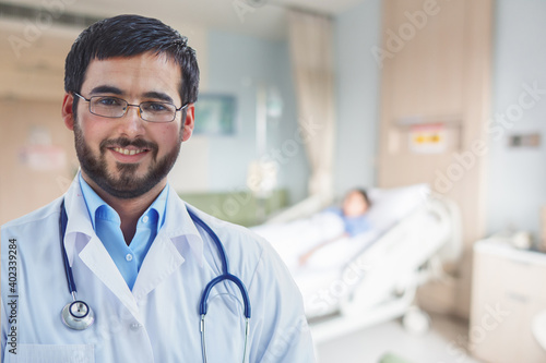 Portrait of smiling Arabian doctor looking at camera with a blurred background of a patient on the bed.