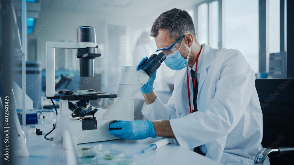 Medical Development Laboratory: Scientist Wearing Face Mask Looking Under Microscope. He Mix Petri Dish Sample. Specialists Working on Medicine, Biotechnology Research in Advanced Pharma Lab Medical Development Laboratory: Scientist Wearing Face Mask Looking Under Microscope. He Mix Petri Dish Sample. Specialists Working on Medicine, Biotechnology Research in Advanced Pharma Lab